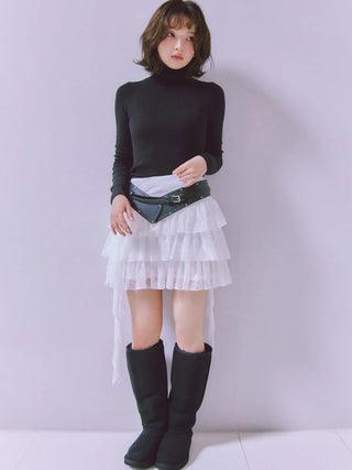 A model stands against a light lavender wall wearing a black long-sleeve mock neck top, a white tiered ruffled mini skirt with a high-low hem, a black faux-leather belt bag, and black boots.