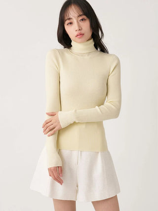 A woman wearing a pale cream ribbed turtleneck sweater and white textured shorts, standing against a plain light background.
