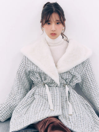 A woman wearing a light gray and white tweed belted coat with a large white faux-fur collar, layered over a white turtleneck sweater and brown pants, posed against a plain white wall.
