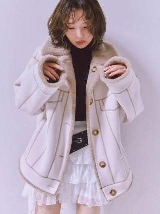 A woman wearing a cream-colored shearling-lined coat with gold buttons, styled over a black turtleneck and a white ruffled skirt, set against a plain light gray background.