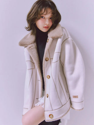 A woman wearing a cream-colored shearling-lined jacket with gold buttons over a black top, posing against a light gray studio background.
