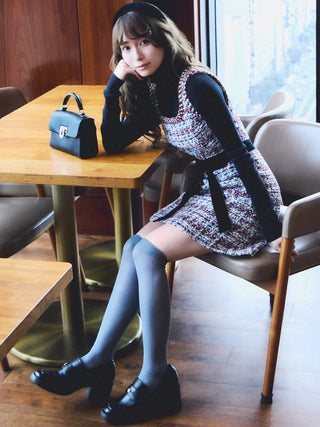 A woman sits on a cafe chair next to a wooden table, wearing a tweed mini dress over a black long-sleeve top, gray striped thigh-high socks, and black loafers. A black handbag rests on the table.