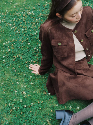 A woman sits on a grassy lawn covered with small white flowers, wearing a brown suede-like jacket and matching skirt over a cream ribbed top. The photo is taken from above at an angle, showing her hand resting on the grass and her legs crossed at the bottom right.