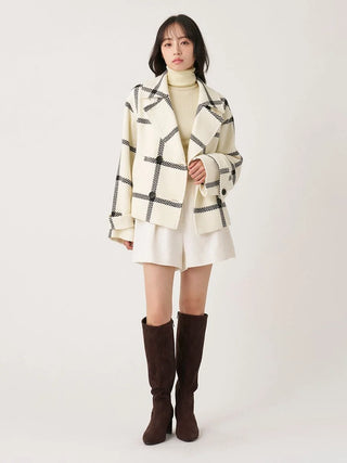 A model stands against a light gray studio background wearing a cream-colored plaid blazer over a cream turtleneck, a cream mini skirt, and dark brown knee-high boots.