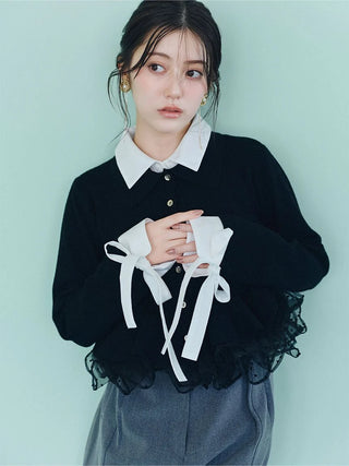 A young woman wearing a black knit long-sleeve top with a white pointed collar and white ribbon tie details at the cuffs, paired with gray trousers, set against a solid pale mint green background.