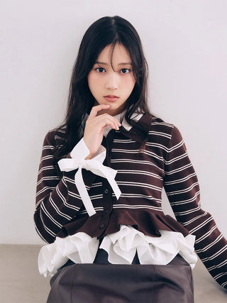 A young woman with long dark hair poses in a brown and white striped long-sleeve blouse with white ruffles and a bow, paired with a dark skirt, against a plain light gray wall.