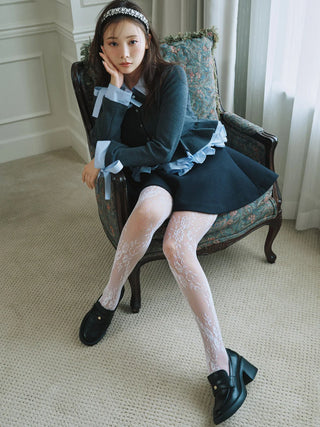 A woman sits in a vintage floral armchair wearing a dark navy blue outfit with light blue ruffle accents, white lace tights, and black loafers in an indoor room setting.