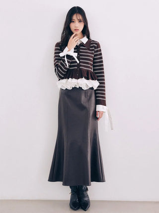 A model stands against a plain light gray wall wearing a long-sleeve brown and white striped knit top with white ruffle trim and a dark brown skirt, paired with black ankle boots.