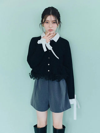 A young woman stands against a pale mint green background wearing a black cardigan with a white collar and bow tie detail, gray shorts, and black knee-high boots.