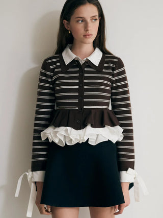 A model wearing a brown and white striped long-sleeve top with a white ruffle hem and white collar, paired with a dark navy mini skirt, standing against a plain off-white wall.