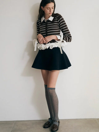 A woman stands against a plain off-white wall wearing a black and white striped long-sleeve top with a white ruffled collar detail, a black pleated mini skirt, sheer gray knee-high socks, and dark brown loafers.
