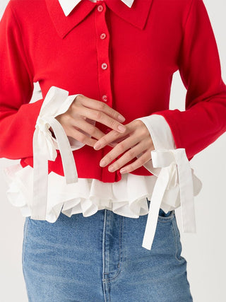 A close-up of a person wearing a bright red long-sleeve button-front top with white ruffled trim and white ribbon bow cuffs, paired with blue jeans against a light neutral background.