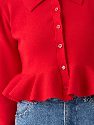 A close-up of a bright red long-sleeve knit top with a pointed collar, button-front closure, and ruffled hem, styled with blue jeans.