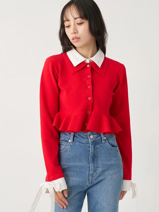 A woman wearing a bright red long-sleeve knit cardigan with a white pointed collar, matching white ruffled cuffs, and blue jeans, photographed against a plain light gray studio background.