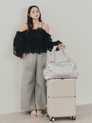 A woman stands indoors next to a cream-colored rolling suitcase, holding a light silver-gray duffel bag on top. She is wearing a black off-the-shoulder ruffled top, wide-leg gray pants, and strappy sandals against a plain light gray wall.