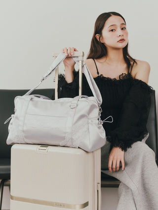 A woman sits on a chair next to a cream-colored suitcase, holding a large light gray duffel bag with lace trim. She is wearing a black off-the-shoulder lace top and a gray skirt.