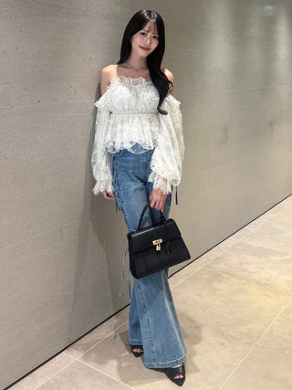 A woman stands indoors against a textured gray wall wearing a white off-the-shoulder lace blouse with long sheer sleeves, blue flare jeans, and black pointed shoes, while carrying a black handbag.