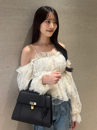 A woman wearing a cream off-the-shoulder lace blouse and blue jeans, holding a structured black handbag with a gold lock against a textured gray wall.