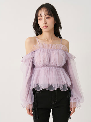 A woman modeling a light purple off-the-shoulder sheer tulle top with long balloon sleeves and a ruffled peplum waist, paired with black pants, against a plain light background.