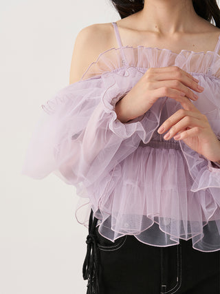 A close-up of a model wearing a light lavender off-the-shoulder tulle top with layered ruffles and puffed sleeves, paired with black pants against a plain light background.
