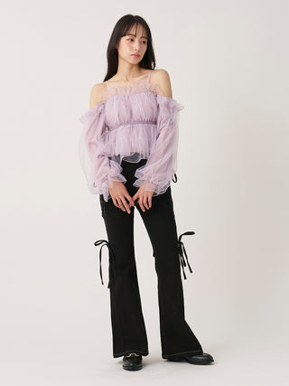 A model stands against a light gray studio background wearing a sheer lavender off-the-shoulder ruffled top paired with black flared pants featuring side tie details and black shoes.