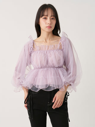 A woman modeling a light lavender puff-sleeve blouse with a ruffled square neckline and sheer tulle layers, paired with black pants, against a plain light gray studio background.