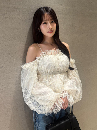 A woman wearing a cream off-the-shoulder lace top with dramatic balloon sleeves, paired with blue jeans and holding a black handbag, standing against a textured concrete wall.